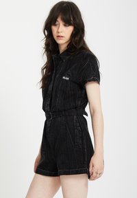 Mono de mezclilla negra con mangas cortas, cintura ceñida con cinturón y un acabado texturizado con líneas sutiles. Incluye un bolsillo lateral con el logo de la marca.