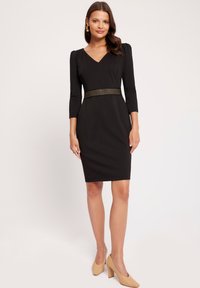 Czarna dopasowana sukienka z rękawami o długości trzech czwartych, dekoltem w serek i zdobionym paskiem w talii. Noszona z cielistymi szpilkami.
