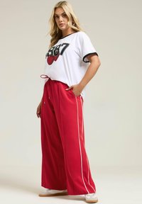 Weißes T-Shirt mit schwarzen Akzenten und grafischem Druck, kombiniert mit roten, weit geschnittenen Hosen mit Kordelzug und weißen Seitenstreifen.