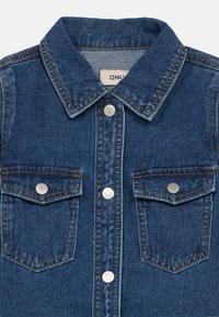 camisa de ganga com colarinho, dois bolsos no peito, botões de pressão prateados, cor azul e detalhes visíveis de costura. Tecido texturizado.