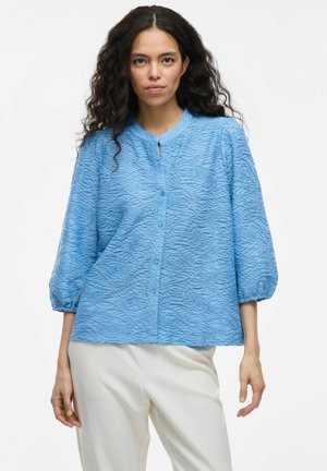 Blouse bleu clair avec un motif de vagues texturé, manches 3/4 et une petite ouverture en forme de clé au niveau du cou. Fermée par des boutons sur le devant.