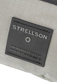 Tessuto grigio con texture a rete, caratterizzato da un'etichetta in pelle nera con il nome del marchio e le coordinate, accentuata da cuciture.