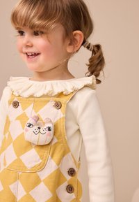 Salopette à carreaux jaune et blanc avec des boutons en bois, portée sur un chemisier crème à manches longues à volants, agrémentée d'un accent en peluche animale.