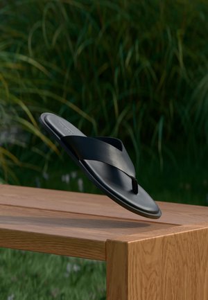 Čierna kožená sandála slide s prekríženými remienkami vznášajúca sa nad drevenou lavičkou vonku s pozadím zelenej vegetácie.