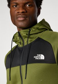 Groene en zwarte ritssluiting jas met capuchon. Heeft een gladde textuur, elastische trekkoorden en een wit logo op de borst.