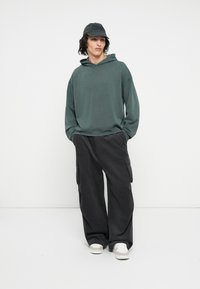 Mørkegrøn hættetrøje med lange ærmer, parret med løstsiddende sorte cargo-bukser med sidelommer. Hvide sneakers fuldender looket.