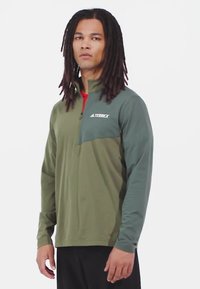 Camicia verde a maniche lunghe con colletto a mezza zip, dotata di un pannello toracico verde scuro, rifiniture rosse e logo "TERREX" in bianco.