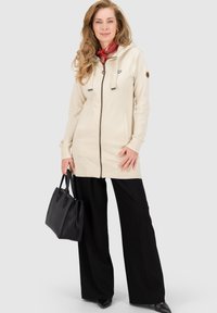 Beige Zip-up-Mantel mit Kapuze, ausgestattet mit Kordelzügen, einem Herz-Logo und grauen Akzenten. Kombiniert mit schwarzen weit geschnittenen Hosen und einer schwarzen Handtasche.