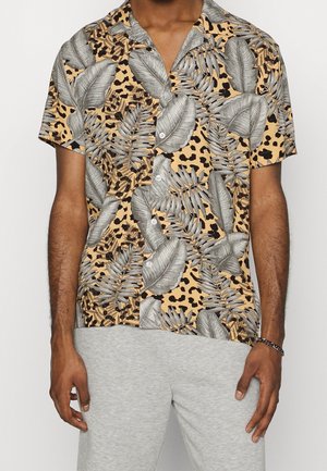 Uomo che indossa una camicia a maniche corte con stampa leopardata beige e nera e grandi foglie tropicali grigie, abbinata a pantaloni grigio chiaro.