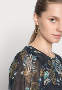 Blusa floral com base azul escura, apresentando padrões verdes e azuis. O modelo usa grandes brincos de argola dourada. Tecido suave e leve, com decote redondo.