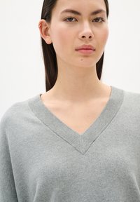 Pull gris à col en V avec une texture douce et des finitions côtelées, porté par un modèle aux cheveux foncés, sur fond blanc uni.