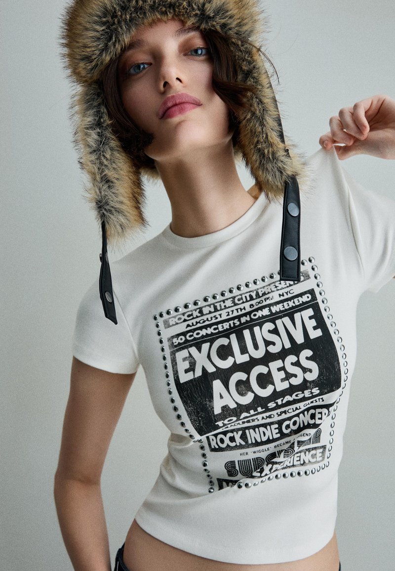 Fehér grafikai póló, fekete nyomtatott mintával, szöveggel és dekoratív szegecsekkel. Szőrmebéléses fülvédős sapka barna részletekkel.