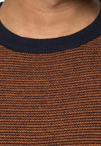 Den stickade tröjan har en texturerad orange kropp med horisontella ränder och en mörkblå ribbad halsringning. Tyget ser mjukt och varmt ut.