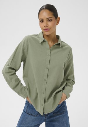 Žena s tamnom kosom vezanom unatrag, odjevena u svijetlozelenu košulju na kopčanje i plave traperice, stoji s rukama u džepovima na jednobojnoj pozadini.