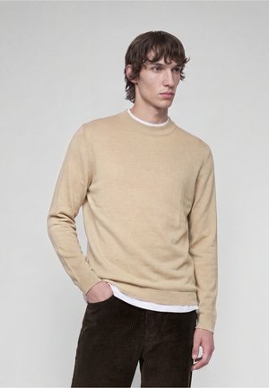 Junger Mann mit braunem, welligem Haar, der einen beigen Pullover über einem weißen Hemd und dunkelbraunen Cordhosen trägt, steht mit einer Hand in der Tasche.