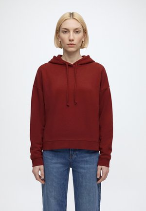 Blonde Frau, die goldene Creolen, einen roten Hoodie und blaue Jeans trägt, steht vor einem schlichten weißen Hintergrund und blickt nach vorne.