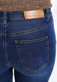 Jeans de mezclilla en azul oscuro con una textura suave. Presenta una parche de cuero con el logotipo "Noisy May" y un bolsillo trasero con costuras contrastantes.
