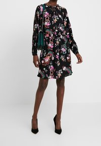 Černé květinové šaty s dlouhými rukávy, stáhnutým pasem a šňůrkovým výstřihem. Vyznačují se živými růžovými a zelenými květinovými vzory. Doplněno černými podpatky.