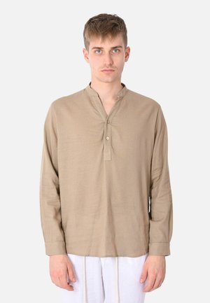 Giovane uomo con capelli corti che indossa una camicia beige a maniche lunghe con patta parziale con bottoni e pantaloni bianchi, in piedi davanti a uno sfondo semplice.