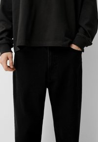 Črna majica z dolgimi rokavi, prekrita preko črnih džins hlač. Hlače imajo sproščen kroj, stranske žepe in subtilne detajle stikanja.