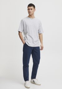 Camiseta gris de manga corta, pantalones cargo azul marino con bolsillos y zapatillas blancas. El modelo está de pie de manera casual con una mano en el bolsillo.
