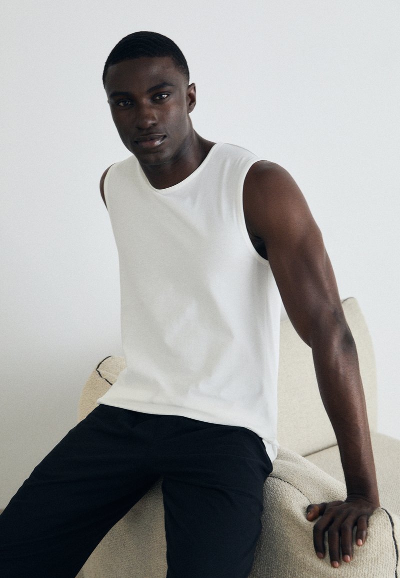 White sleeveless shirt in soft cotton fabric, paired with dark pants. Model seated on a textured light-colored chair, neutral background.