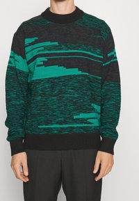 Pull en tricot avec base noire et motifs de rayures abstraites vertes, col et poignets côtelés, coupe décontractée. Texture douce.