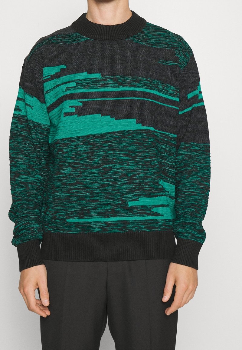 Pull en tricot avec base noire et motifs de rayures abstraites vertes, col et poignets côtelés, coupe décontractée. Texture douce.