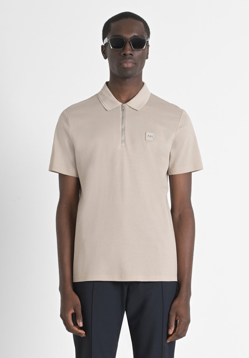 Polo de color beige claro con mangas cortas, cierre de cremallera y un parche de logo cuadrado en el pecho. El modelo lleva gafas de sol negras.