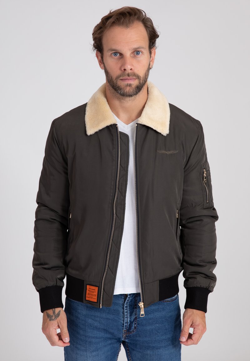 Ciemnozielona kurtka bombowiec z beżowym kołnierzem z sztucznego futra, z zapięciem na zamek z przodu, ściągaczami na mankietach oraz naszywką na dolnym brzegu.