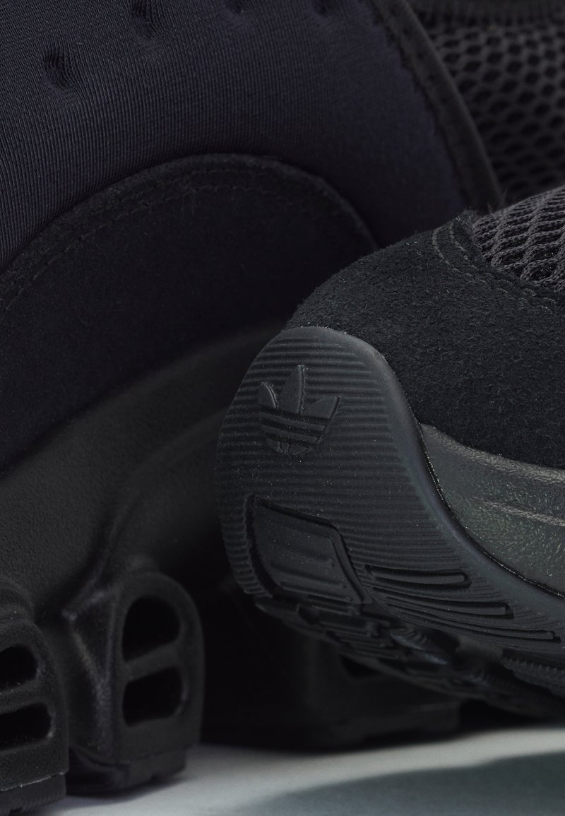 Chaussures de sport noires avec un mélange de matériaux en maille et en daim, une semelle texturée et un logo sur le talon. Vue rapprochée mettant en avant les détails.