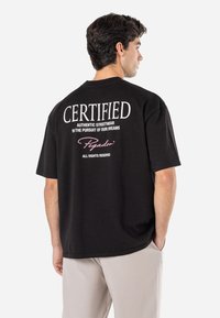 Juodas medvilninis marškinėlis su apvalia iškirpte, laisvas kirpimas, su baltais ir rožiniais užrašais gale: "CERTIFIED Authentic Streetwear Pegador."