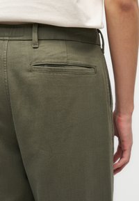 Κοντινό πλάνο στη μέση ενός ατόμου που φορά ελιάς πράσινο παντελόνι με πίσω τσέπη με φάσα και ένα λευκό πουκάμισο μέσα στην μέση.