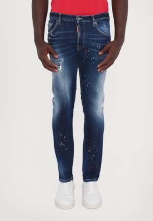 Donkerblauwe versleten denim jeans met verfspatten, met een slim fit, vijf zakken en een knoopsluiting. Witte sneakers zichtbaar.