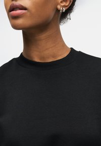 Κοντινό πλάνο ενός ατόμου που φορά μικρούς ασημένιους κρίκους στα αυτιά και μαύρο μπλουζάκι με λαιμόκοψη, εμφανίζοντας τα χείλη, το λαιμό και μέρος του αυτιού.