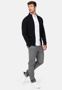 Cardigan en tricot navy par-dessus une chemise blanche avec des boutons noirs, associé à un pantalon gris clair et des baskets blanches, présentant un détail de poche zippée.
