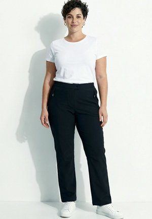 Mujer con cabello corto y rizado de pie sobre fondo blanco, lleva una camiseta blanca, pantalones negros con bolsillos con cremallera y zapatillas blancas.