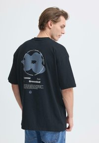 T-shirt noir oversize avec un motif graphique au dos, comprenant du texte et une forme arrondie. En coton. Vue de face non visible.