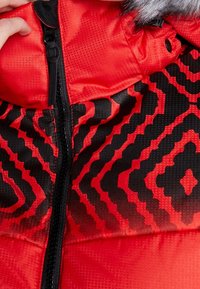 Veste d'hiver rouge avec motif géométrique noir, fermeture éclair noire centrale et capuche doublée de fourrure, tenue à la main près du haut.
