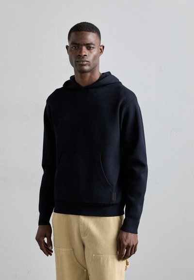 Hoodie en coton navy avec une poche avant, des poignets côtelés et une coupe décontractée, associé à un pantalon beige avec des poches latérales et des boutons.