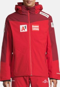 Rote wasserdichte Jacke mit Kapuze, mit Logos auf der Brust und den Ärmeln, Reißverschluss und einer leicht strukturierten Oberfläche.