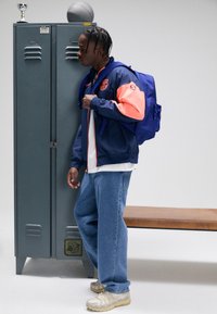 Marinblå och korallfärgad jacka med logotyp, vit skjorta, blå jeans och ljusgrå sneakers. Blå ryggsäck. Metallskåp och träbänk i bakgrunden.