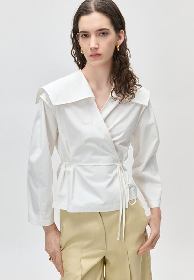 Kobieta o kręconych brązowych włosach, ubrana w biały, kopertowy top z dużym kołnierzem, beżowe spodnie, złote kolczyki koła i złoty pierścionek, stojąca na białym tle.