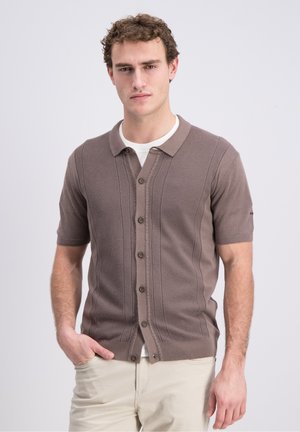 Young man wearing a brown short-sleeve buttoned cardigan over a white t-shirt, with beige pants, standing against a light neutral background.