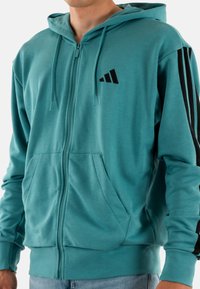 Felpa con zip color teal, dotata di due tasche frontali, strisce nere sulle maniche e un logo nero. Realizzata in morbido tessuto testurizzato e con cappuccio.