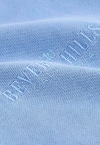 Tessuto azzurro chiaro con superficie strutturata, caratterizzato da testo in rilievo "BEVERLY HILLS CALIFORNIA" in un azzurro più chiaro. Design semplice e finitura liscia.