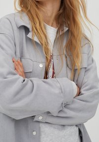 Lys grå, tekstureret jakke med knaplukning, to brystlommer og en afslappet pasform, båret over en grå sweatshirt.