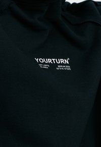 Polo negro de algodón con cuello alto, que presenta una textura acanalada y un logotipo impreso en blanco. Incluye "YOURTURN", "Berlín 2023" y coordenadas geográficas.