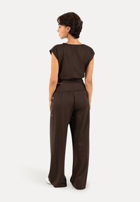 Femme debout de dos portant un haut sans manches marron foncé et un pantalon à jambes larges avec des chaussures blanches sur un fond uni.