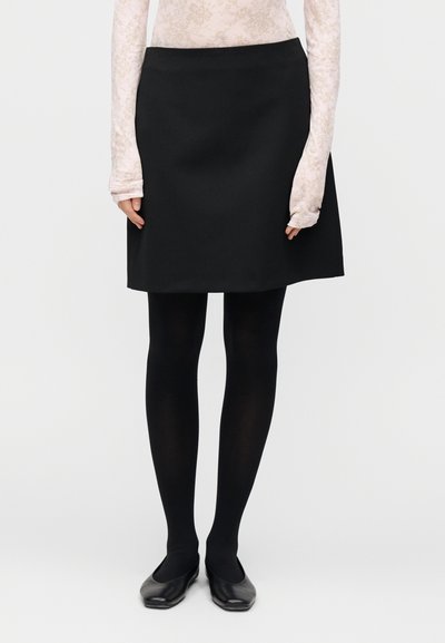 Jupe trapèze noire en tissu lisse, associée à des collants opaques noirs et des chaussures plates noires. Haut à manches longues à motif floral visible en dessous.